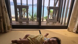 A child lying on a tatami mat with a view of the sea and chairs outside a window. - Amami Lucky House (Amamishi,Kasaricho,OoagiKiseuttabaru)