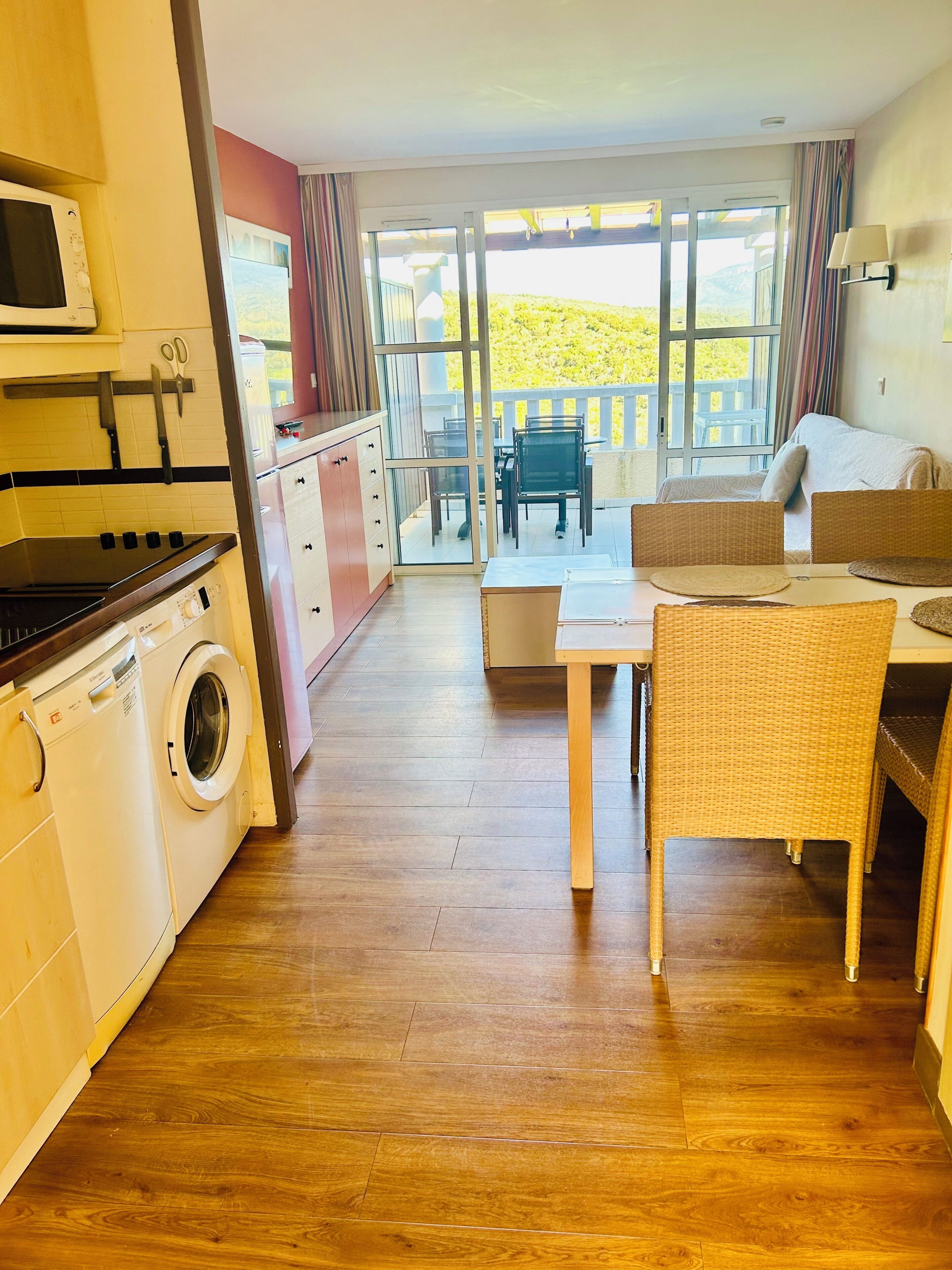 A compact kitchen with a washing machine, a dining area with wicker chairs, and a balcony with a view.