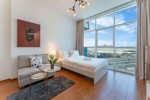 A modern hotel room with a large window, a sofa, a bed, a small table with a plant, and a painting on the wall. - Bright and Spacious Studio in JVT l Free Pool,Gym (Jumeirah Village Triangle)