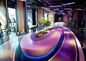 A modern gym with exercise equipment, a curved seating area, and a plant centerpiece. - Bright and Spacious Studio in JVT l Free Pool,Gym (Jumeirah Village Triangle)
