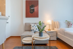 A modern living room with a sofa, a small table with a plant, a lamp, and a painting on the wall. - Bright and Spacious Studio in JVT l Free Pool,Gym (Jumeirah Village Triangle)
