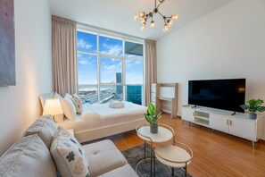 A modern living room with a large window, a sofa, a TV, and a small table with a plant. - Bright and Spacious Studio in JVT l Free Pool,Gym (Jumeirah Village Triangle)