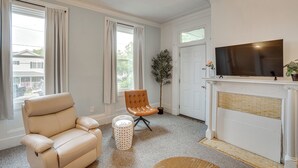 Een woonkamer met een beige leren bank, een klein rond voetenbankje, een potplant, een open haard en een televisie.