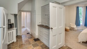 Een kamer met kitchenette, een deur naar een andere kamer, een bed en een raam met gordijnen.