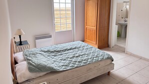 Une chambre à coucher avec un lit, une armoire en bois, une fenêtre et une salle de bain visible à travers une porte ouverte.