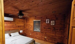 Ein Schlafzimmer einer Holzhütte mit Bett, Deckenventilator, Klimaanlage und Fenster mit Jalousien.