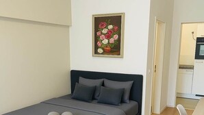 Un dormitorio moderno con una cama, almohadas, un cuadro y una kitchenette al fondo.