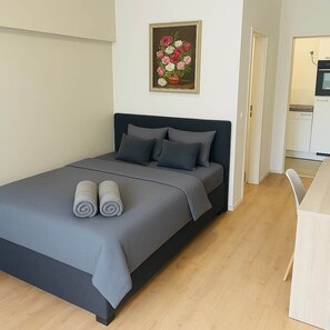 Een moderne slaapkamer met een bed, kussens, een schilderij en een kitchenette op de achtergrond.