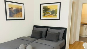 Una cama bien hecha con ropa de cama gris y dos toallas enrolladas, un cabecero negro y dos cuadros en la pared.