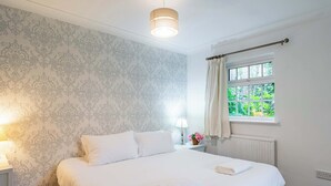 A neatly made bed with white linens, a bedside lamp, and a window with curtains. - 2 bed Bungalow in Towcester by ARP Homes, 15 min from Silverstone, Free Parking, Perfect for Families and Contractors (West Northamptonshire)