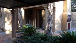 Un espacio al aire libre techado, con columnas, un banco y un sendero que lleva a la entrada de un edificio.