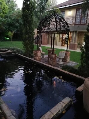 A pond with a gazebo, brick seating, and a house in the background. - Giardino a 194 (Welkom)