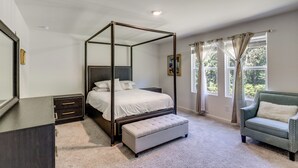 Uma cama de dossel com um banco combinando, uma mesa de cabeceira em madeira escura, uma poltrona verde e uma janela grande com cortinas.