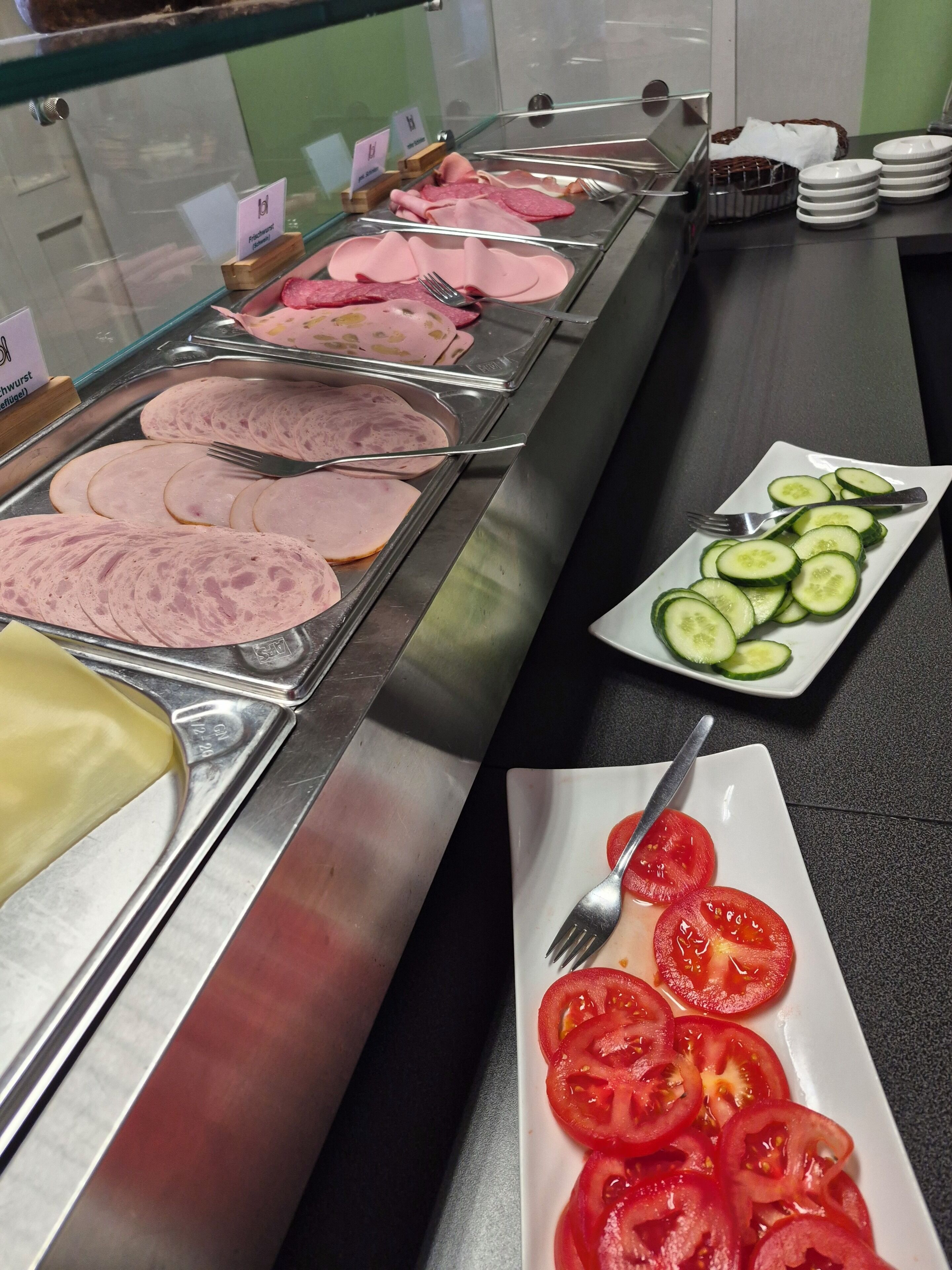 A buffet with various sliced meats and a plate of sliced cucumbers and tomatoes.
