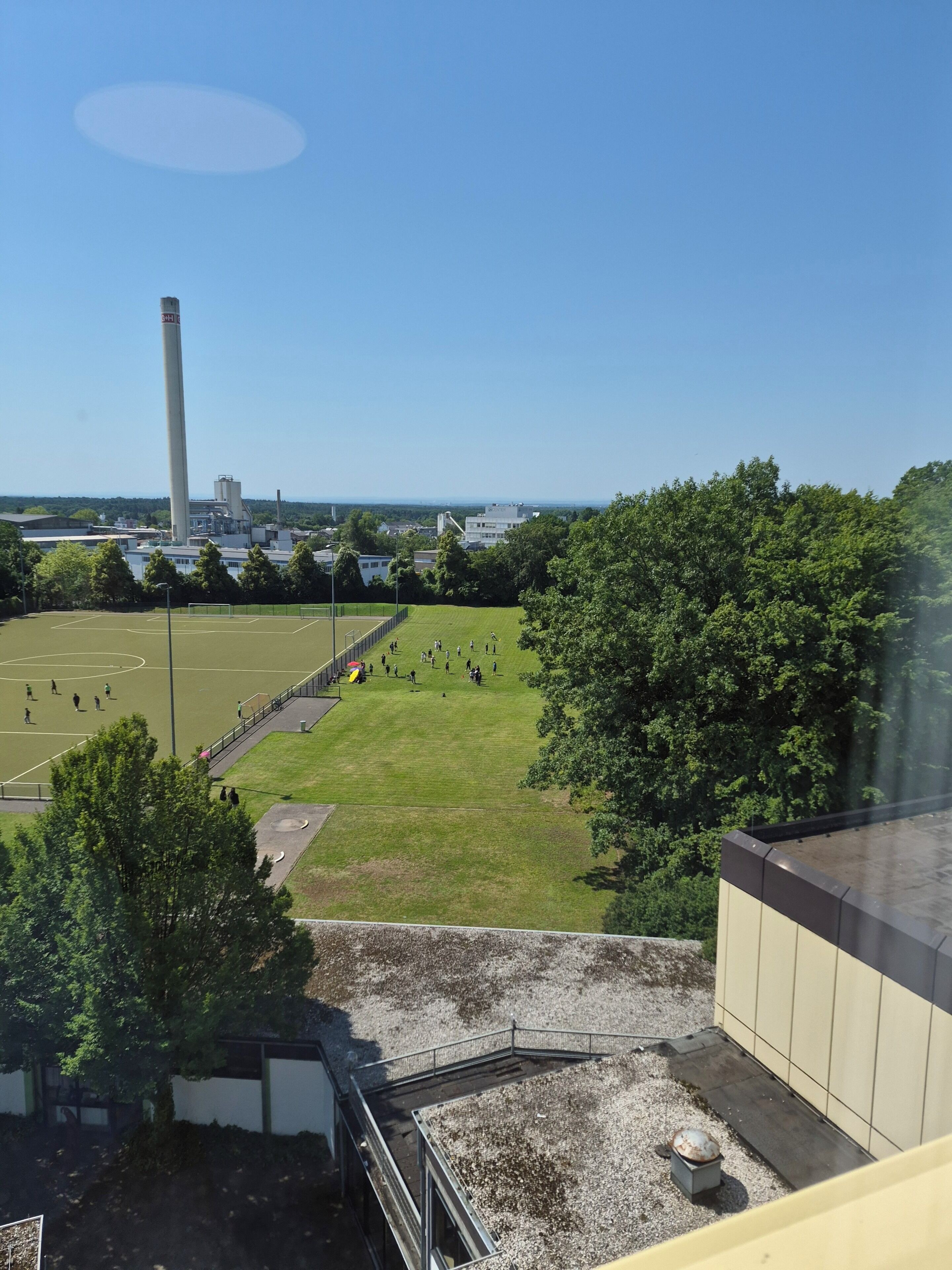 A view of a sports field with people playing, a tall chimney, and industrial buildings in the background.