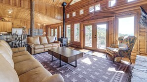 A spacious living area with a sofa, a coffee table, and a patterned rug. There is a fireplace, a dining area with chairs, and a large window offering a view of the outdoors. - Hot Tub + Fire Pit: Spacious Waynesville Escape! (Waynesville)