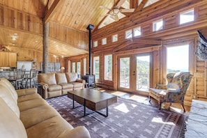A spacious living area with a sofa, a coffee table, and a patterned rug. There is a fireplace, a dining area with chairs, and a large window offering a view of the outdoors. - Hot Tub + Fire Pit: Spacious Waynesville Escape! (Waynesville)