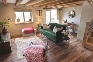 Ein gemütliches Wohnzimmer mit einem grünen Sofa, einem weißen Hocker, einem gemusterten Sessel und einem Couchtisch aus Holz.