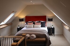 Ein Schlafzimmer mit schrägen Wänden, einem Bett mit gemusterten Kissen, Nachttischen mit Lampen und einer Holzbank mit Handtüchern.