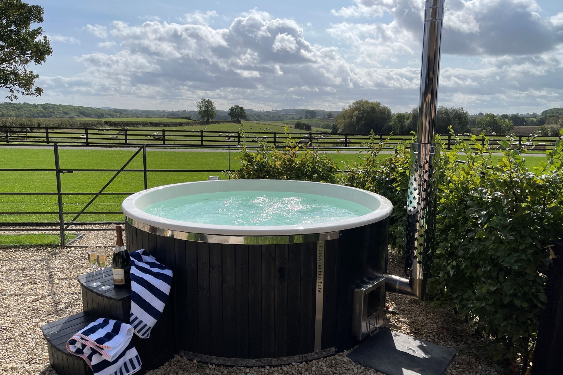 A hot tub with a wooden exterior, surrounded by a gravel area, and a metal fence in the background.