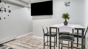 A modern room with a flat-screen TV, a wall clock, a potted plant, and a dining table with chairs.