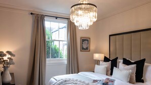 Una camera da letto con un letto grande, un lampadario, una finestra con tende, un comodino con un vaso e un quadro appeso al muro.