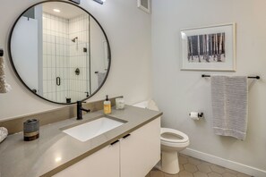 A bathroom with a round mirror, a sink, a toilet, a framed picture on the wall, and a towel rack. - Long-Term Stay in Denver! Dog-Friendly Studio (Denver)