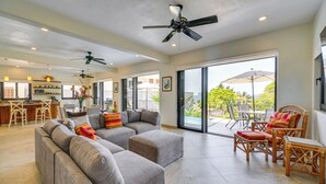 Un vaste espace de vie comprenant un grand canapé d’angle gris, une table à manger et des chaises en osier. On y trouve également des ventilateurs de plafond, un coin cuisine et de grandes portes-fenêtres coulissantes donnant sur un balcon.