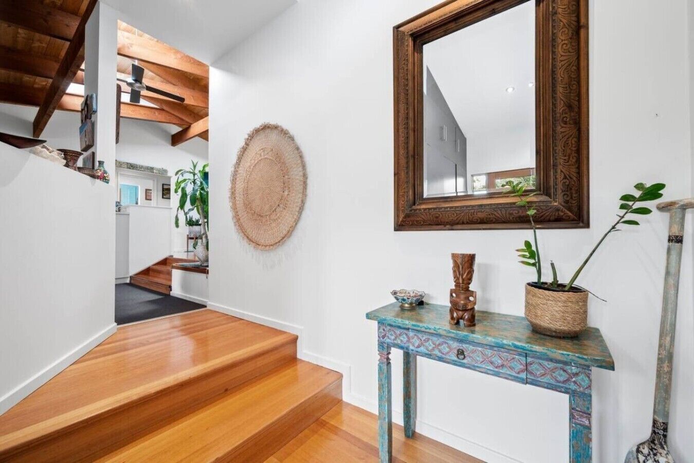 A staircase with wooden steps leading to a landing, a round woven wall hanging, a mirror with an ornate frame, and a small table with a plant and decorative items.