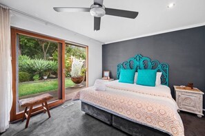 Ein Schlafzimmer mit einem großen Bett, einem Deckenventilator, einem Nachttisch und Blick auf einen Garten durch eine Schiebetür.