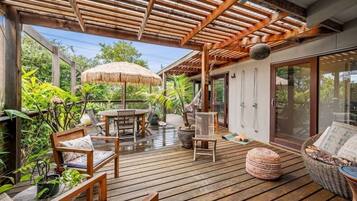 Um deck de madeira com móveis de exterior, um guarda-sol de palha e vasos com plantas.