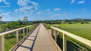 A wooden boardwalk with metal railings, leading through a grassy field with trees and buildings in the background. - Pool Access! Family Getaway in Hilton Head Resort (Hilton Head Island)