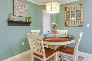 A dining area with a round table, white chairs, a hanging light fixture, a decorative wall piece, and a small shelf with vases. - Pool Access! Family Getaway in Hilton Head Resort (Hilton Head Island)