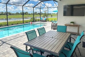 A covered outdoor area with a pool, a table with chairs, and a TV mounted on the wall. - Resort Golf Living: New Home, Elite Club Perks (Lehigh Acres)