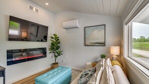 Una sala moderna con un sofá, un reposapiés azul, un televisor montado en la pared, un aire acondicionado y una ventana con vistas.