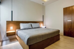 A modern bedroom with a wooden headboard, a bed with a grey quilt, bedside lamps, and a wardrobe. - Modern Condo: Steps to Mamitas Beach Club/5thAve (Playa del Carmen)