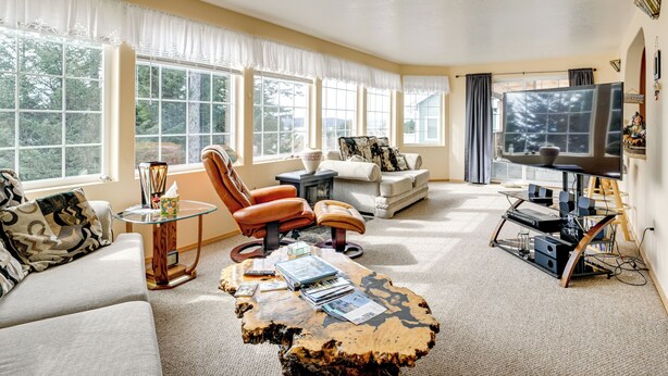 Un salon comprenant un canapé, un fauteuil, un pouf, une table basse, un meuble pour la télévision et de grandes fenêtres.