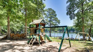 A playground with a wooden playhouse, swings, and a slide, surrounded by trees and a lake. - It's Our Pleasure! Lake Escape w/ Boat Ramp (Macon)