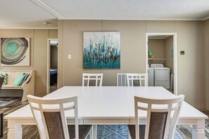Un coin repas avec une table et des chaises blanches, un tableau au mur et, au fond, un espace dédié à la buanderie.