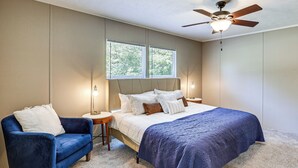 A bedroom with a bed, a blue armchair, two bedside lamps, a ceiling fan, and two windows. - It's Our Pleasure! Lake Escape w/ Boat Ramp (Macon)