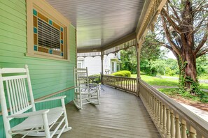 A porch with a rocking chair and a table, a tree, and a view of a green lawn and other houses.