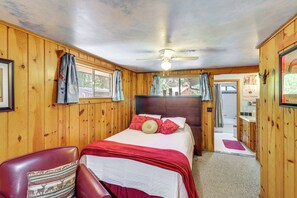 Un dormitorio de cabaña de madera con una cama, una silla, un ventilador de techo y un baño visible a través de una puerta abierta.
