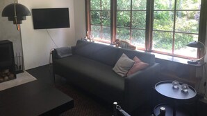 Ett vardagsrum med en soffa, en tv som är monterad på väggen, ett fönster med utsikt över grönområden och ett litet bord med dekorativa föremål.