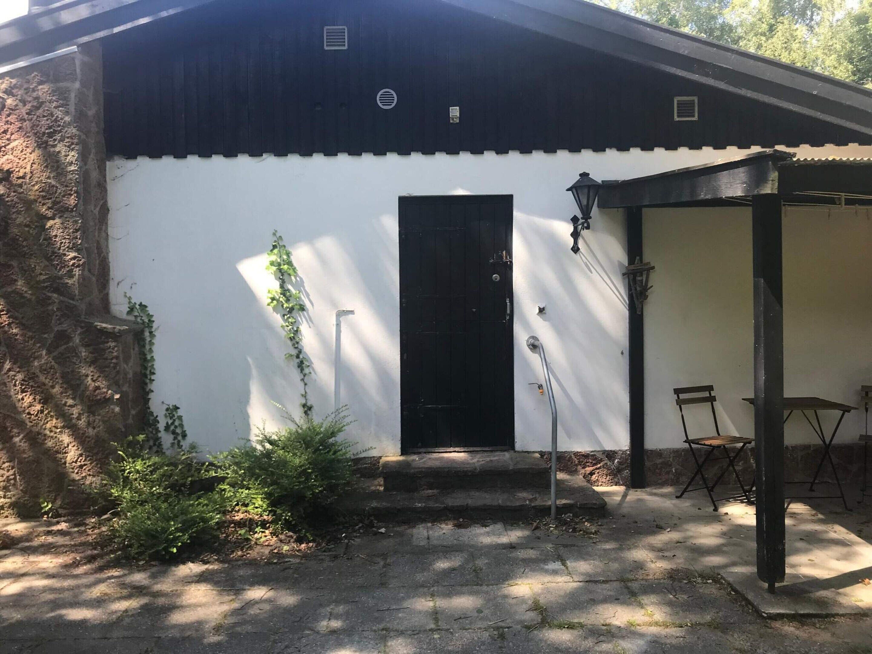 Edificio con puerta negra, zona de patio techada con sillas y pared de piedra con vegetación.