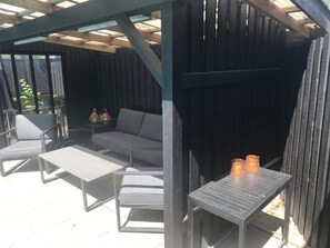 An outdoor patio with a wooden pergola, a grey sofa, two white chairs, and a small table with two candles.