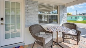 Un patio techado con sillas de mimbre y una mesa, un muro de piedra y una puerta con vidrio esmerilado.
