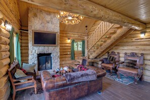 LâintĂ©rieur dâune cabane en rondins, avec une cheminĂ©e en pierre, des meubles en cuir, un escalier et un lustre.