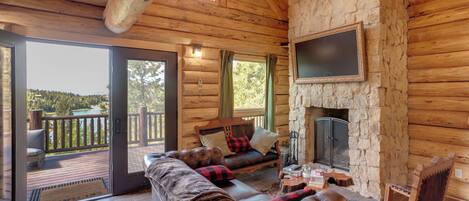 A log cabin interior with a stone fireplace, a leather sofa, and a deck with a view of the outdoors.
