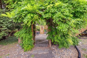 A wooden archway leading to a covered patio area with a brick wall and a wooden fence in the background. - Unique Unit w/ Patio & Grill in Portland Area! (Milwaukie)