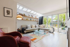 Ein modernes Wohnzimmer mit einer Couch, einem Couchtisch und einem roten Sessel. Regale mit Dekorationselementen und ein großes Fenster mit Blick ins Freie.